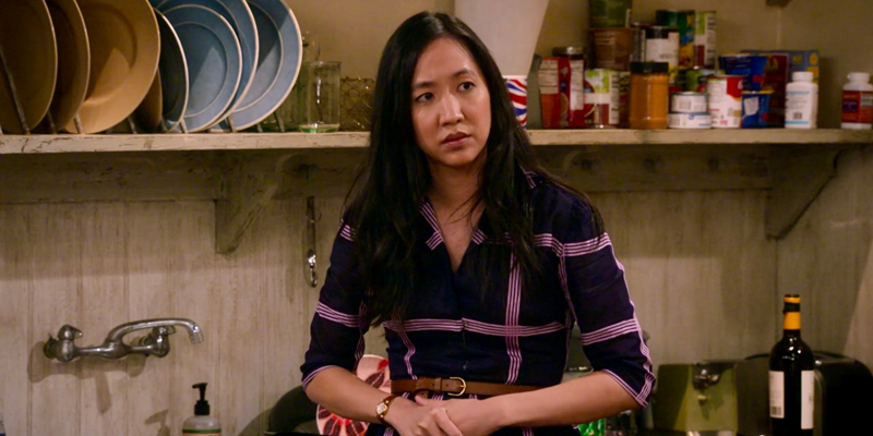 Ellen in How I Met Your Father, an Asian lesbian, is in a flannel purple dress and a confused look on her face in front of a kitchen wall with pots hanging above her head.