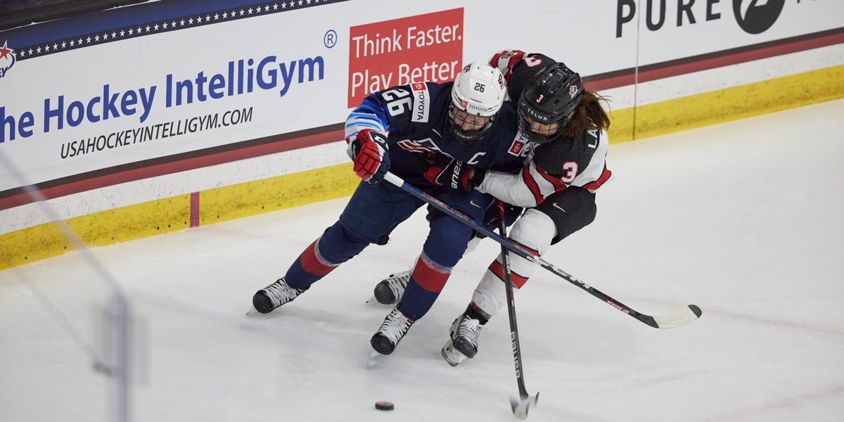 A women's Canadian and American hockey player embrace on the ice as they go for a puck.