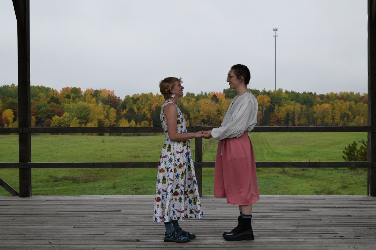 A photo of Erin marrying their spouse in the insect-patterned dress they made. Their spouse is wearing a pink skirt and flowy white top.