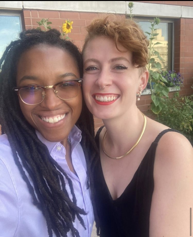 Mae and Nicky pose together smiling. Nicky is Black with a long protective hairstyle. with a blue collared shirt and glasses. Mae is white and has short, curly red hair with shaved sides. Mae is wearing red lipstick, a black dress, and gold earrings and a necklace. Both Nicky and Mae are smiling warmly.