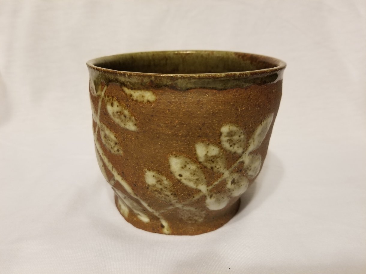 This little ceramic bowl is brown with white a white leaf pattern on it as though a plant were laid on it to create a print.