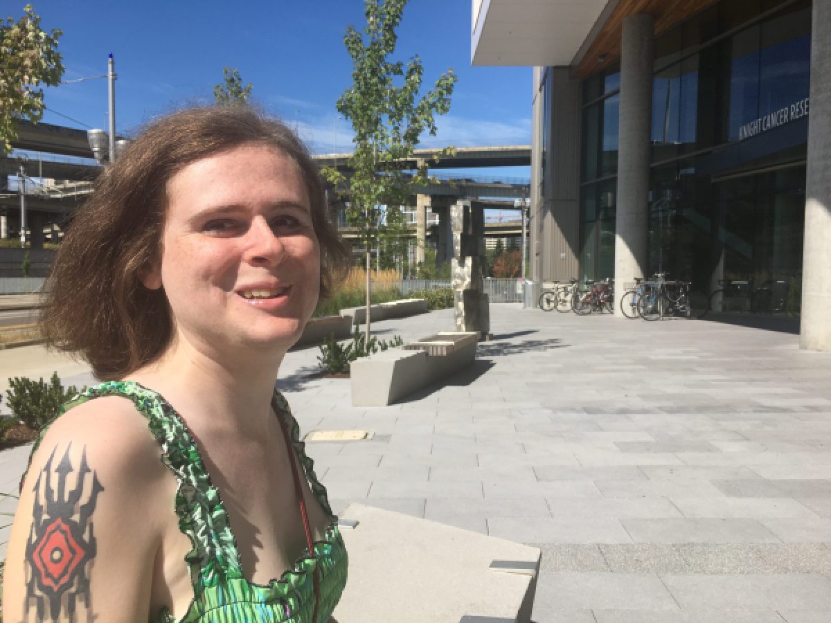 A photo of Wenda smiling in the sunshine. Wenda has pale skin and light reddish brown hair, a tattoo on her upper arm and is wearing a green patterned sundress.