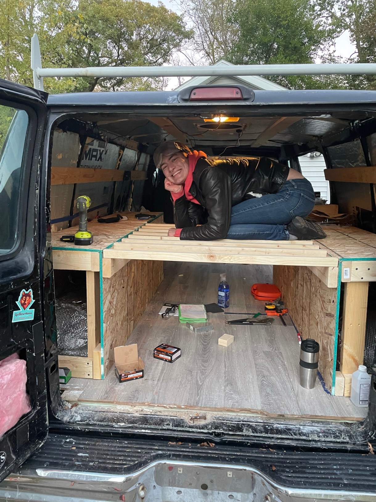 Here, Karalyn poses mid-build of the platform in the van, a drill resting on the wooden platform and the van floor holding tools and a thermos. Karalyn is smiling.