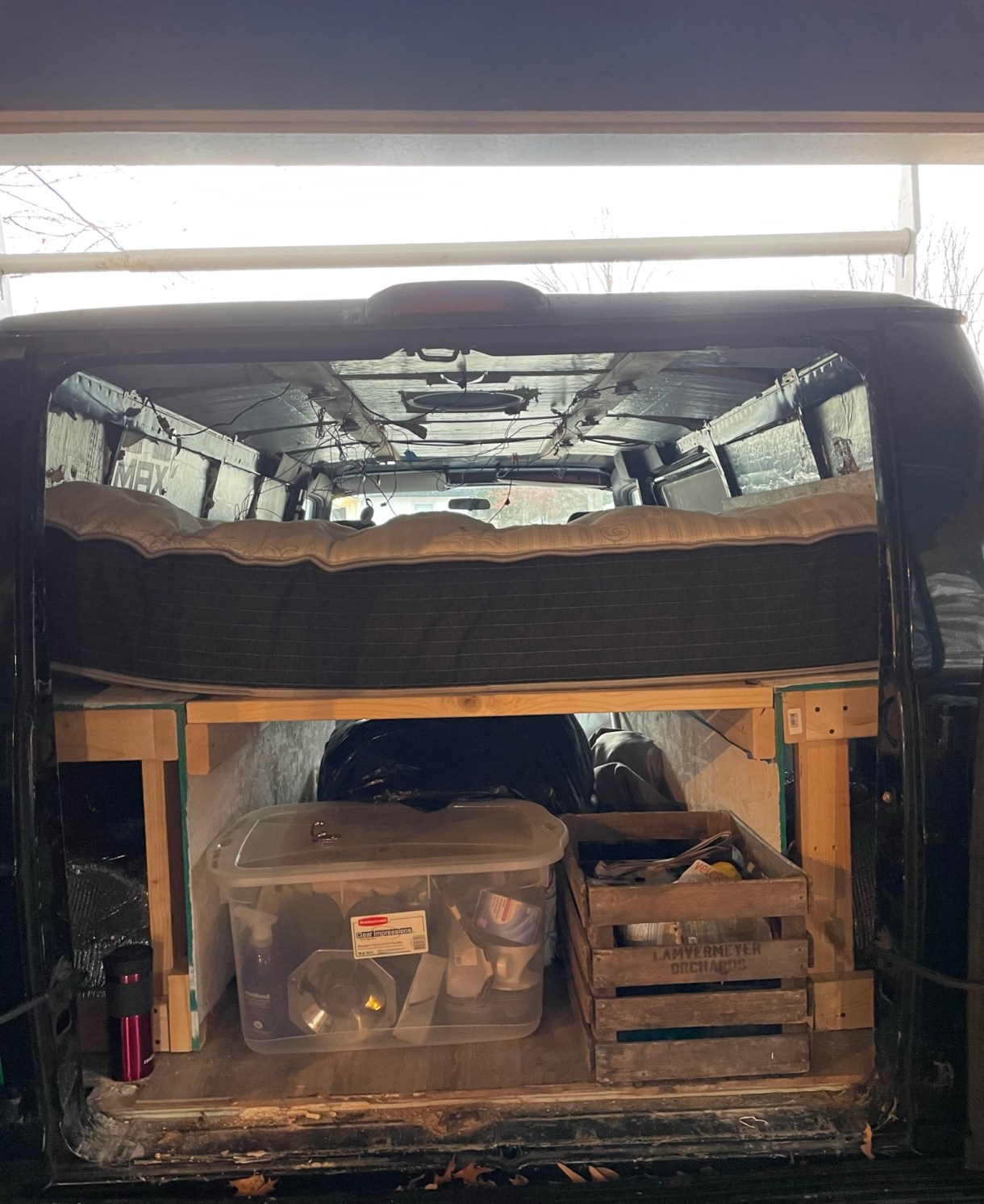 This is a look inside their van. There is a mattress on top of a wooden platform. Underneath are several storage crates and boxes holding supplies and items.