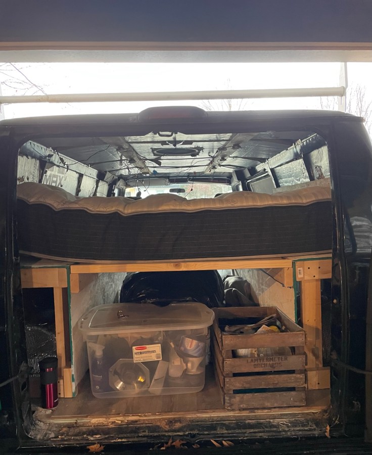 This is a look inside their van. There is a mattress on top of a wooden platform. Underneath are several storage crates and boxes holding supplies and items.