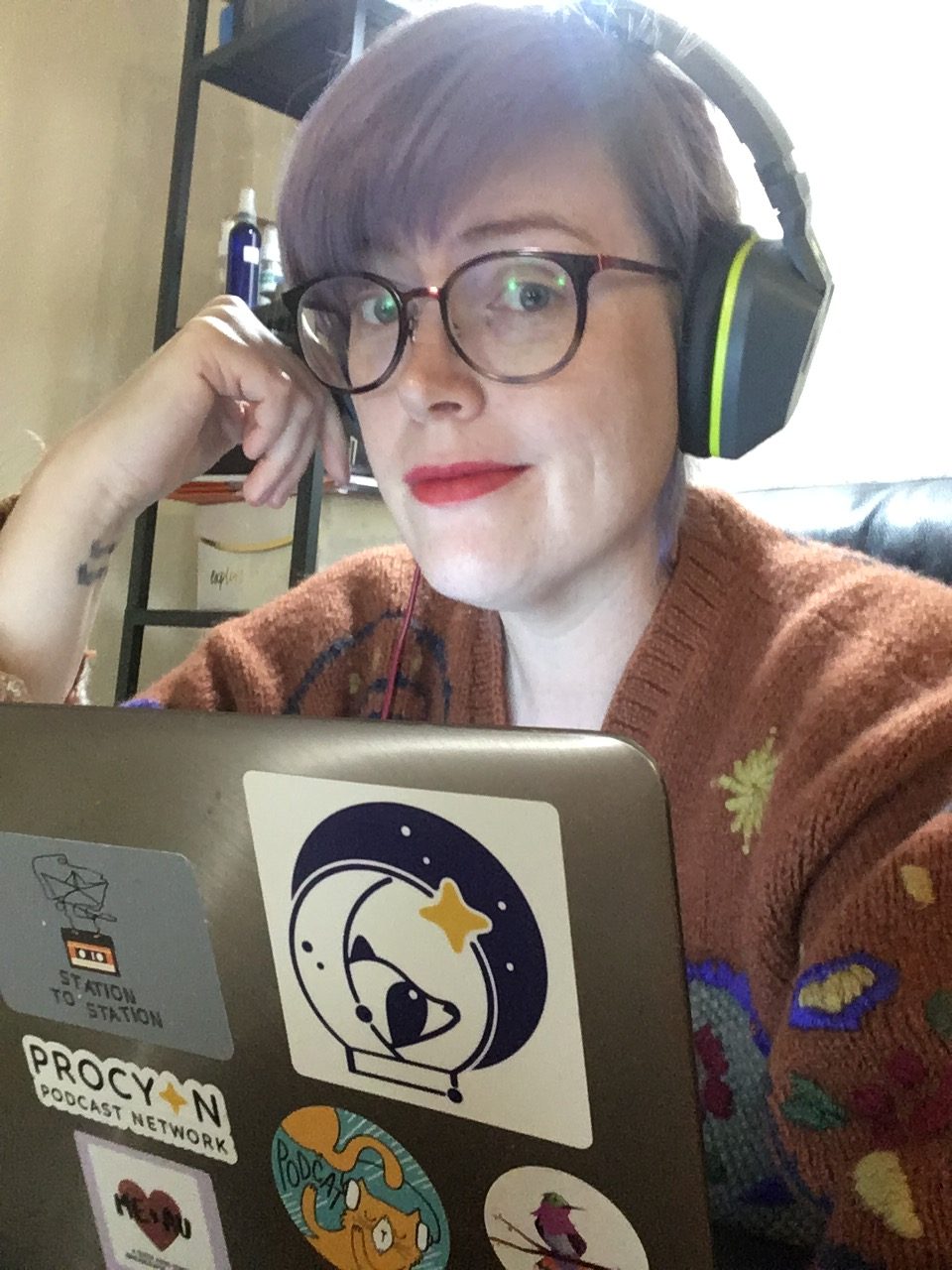 The podcaster, Andrew, looks at the camera while wearing headphones and sitting at a laptop. They have brown hair, light eyes, glasses, and are wearing a sweater.