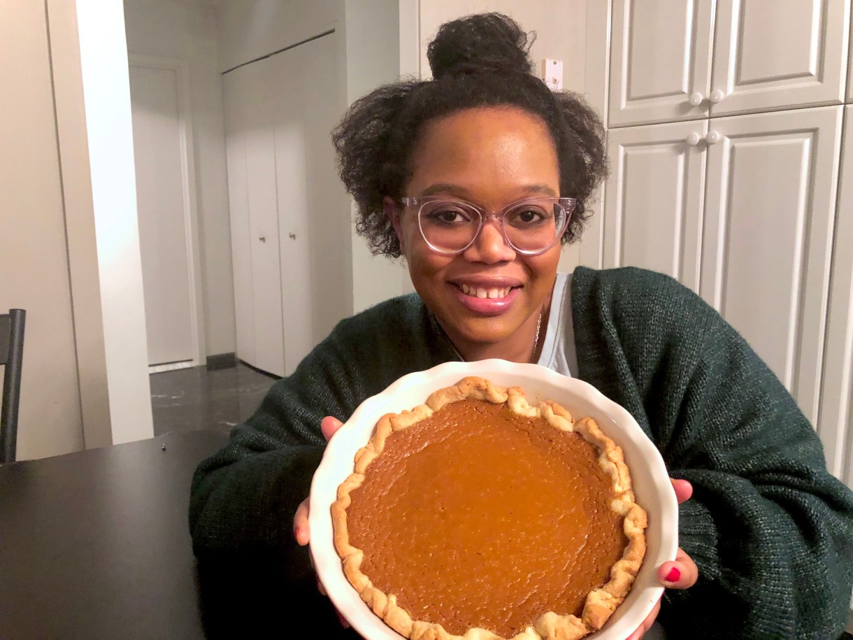 Carmen here smiles and holds a pumpkin pie up! Carmen is a Black woman with her hair up in a partial bun. She is wearing a sweater and clear glasses.