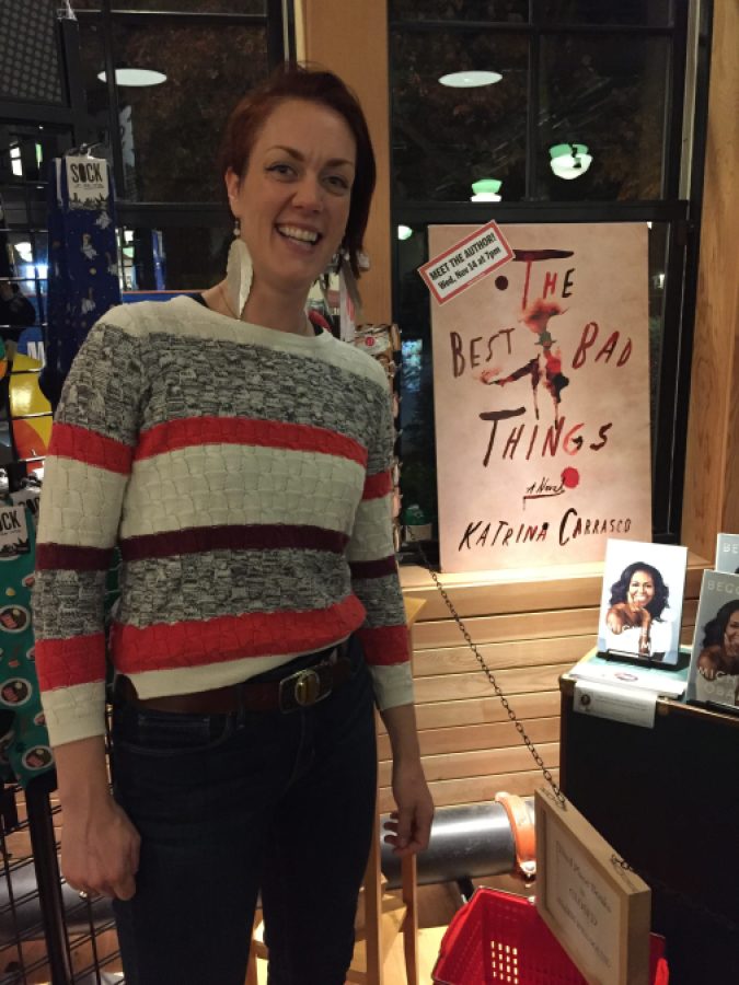 A photo of Katrina at a meet the author event. She is smiling and has a giant cover of her book, The Best Bad Things, behind her. Katrina is a person with pale skin with short brown hair who is wearing a sweater and feather earrings.