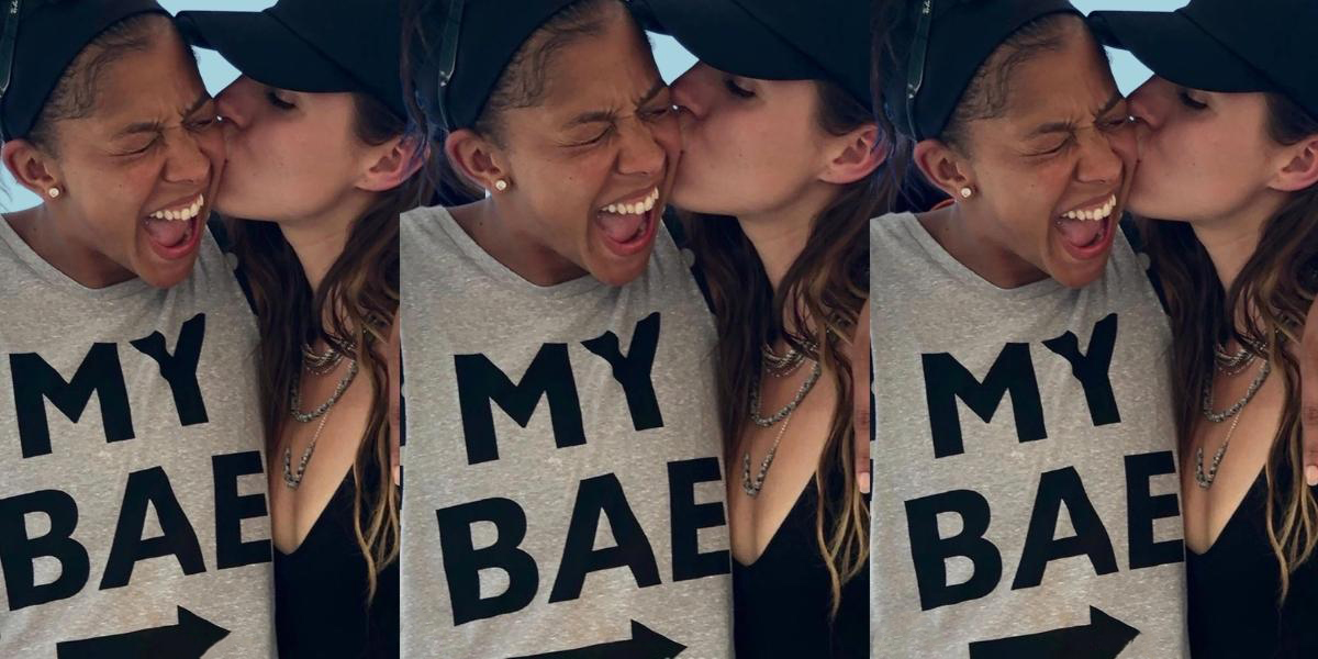A cut out collage of Candace Parker and her wife. Her wife is kissing her cheek while Candace laughs. Candace is wearing a tank top that says "My Bae" that points at her wife.