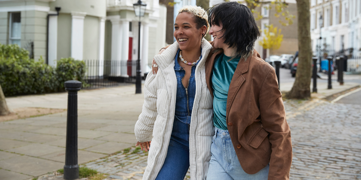 Two women smile with their arms around each other as they walk outside.