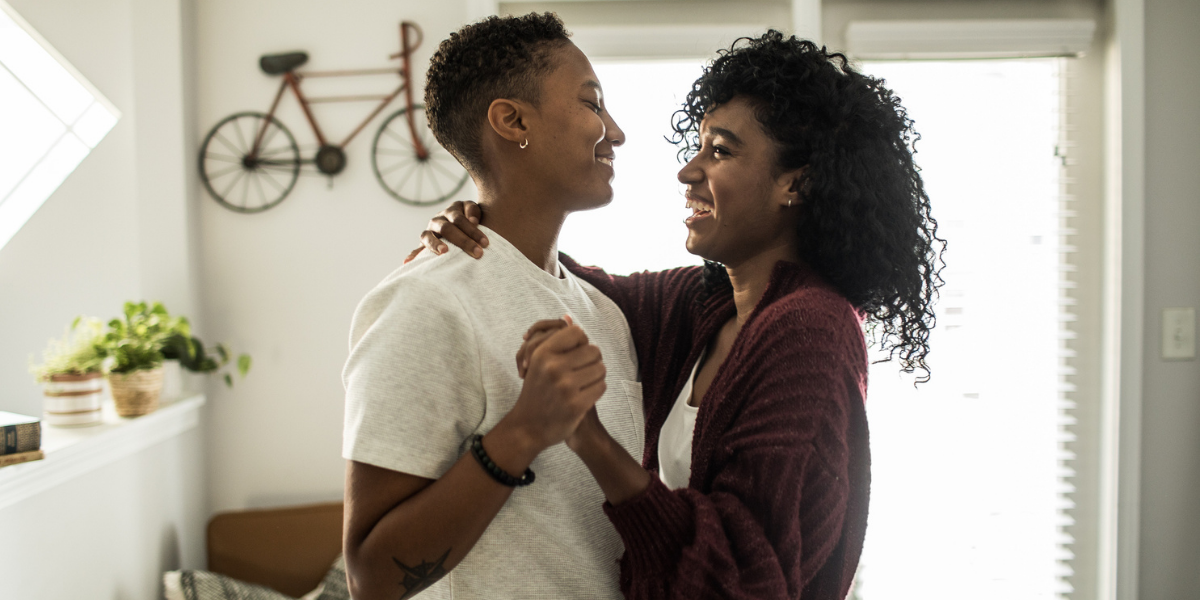 Two Black women dance together while smiling in their home.