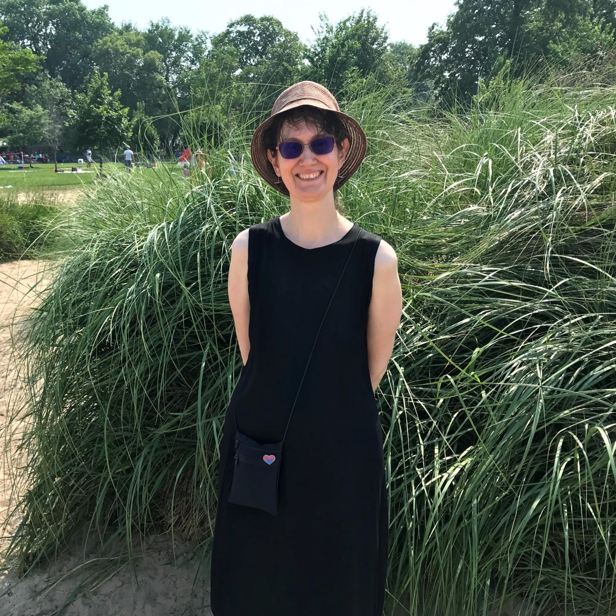 a photo of Cleo smiling in a hat and sunglasses in front of grassy sand dunes in a public park