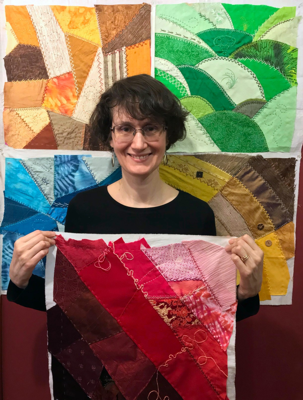 Cleo, a white woman with wavy brown hair and glasses, holds up a basted quilt square in tones of dark brown to burgundy to red to pink. She is smiling.