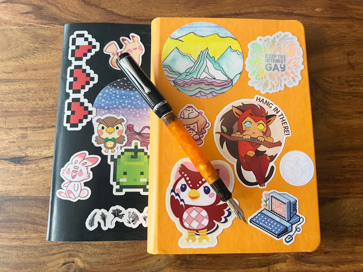 An open fountain pen rests on top of two journals, each covered in stickers. Two of the stickers are even Autostraddle fundraiser stickers!