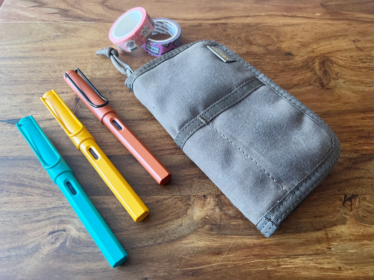 Three fountain pens rest on a wooden table near a carrying pouch and some washi tape.