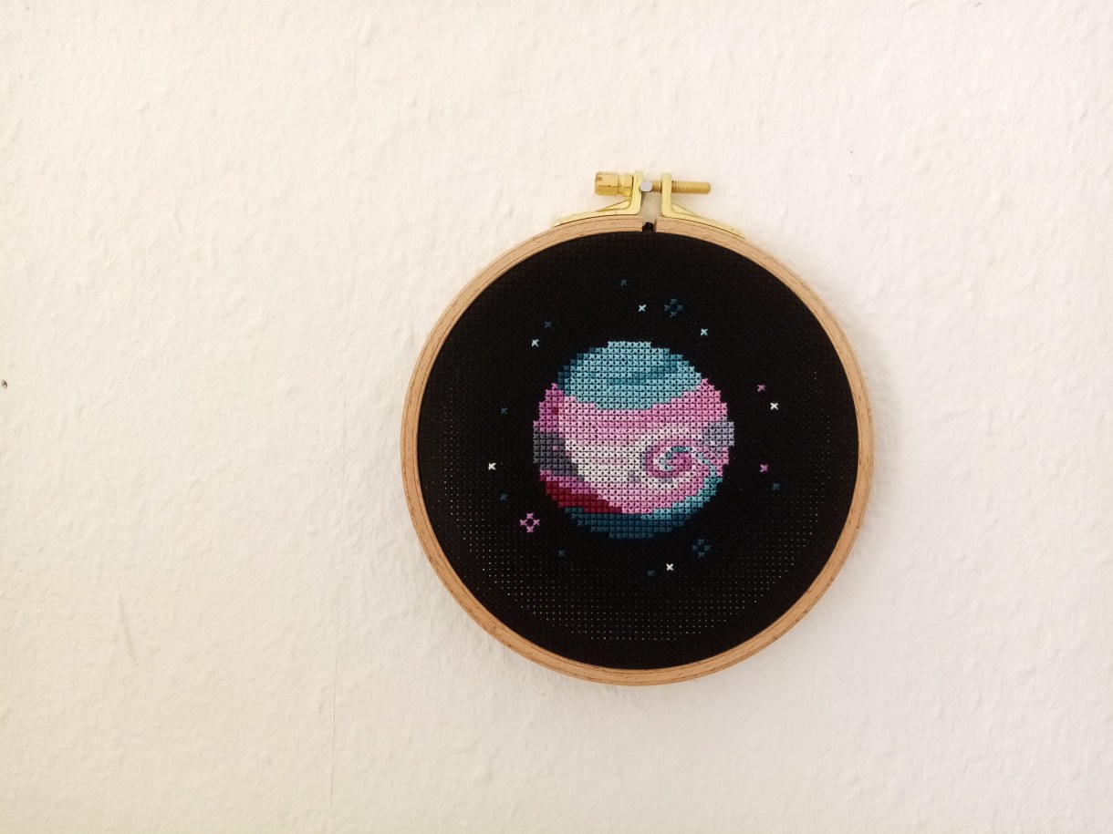 an embroidery piece of a pink, blue and white planet against a black backdrop, within an embroidery hoop