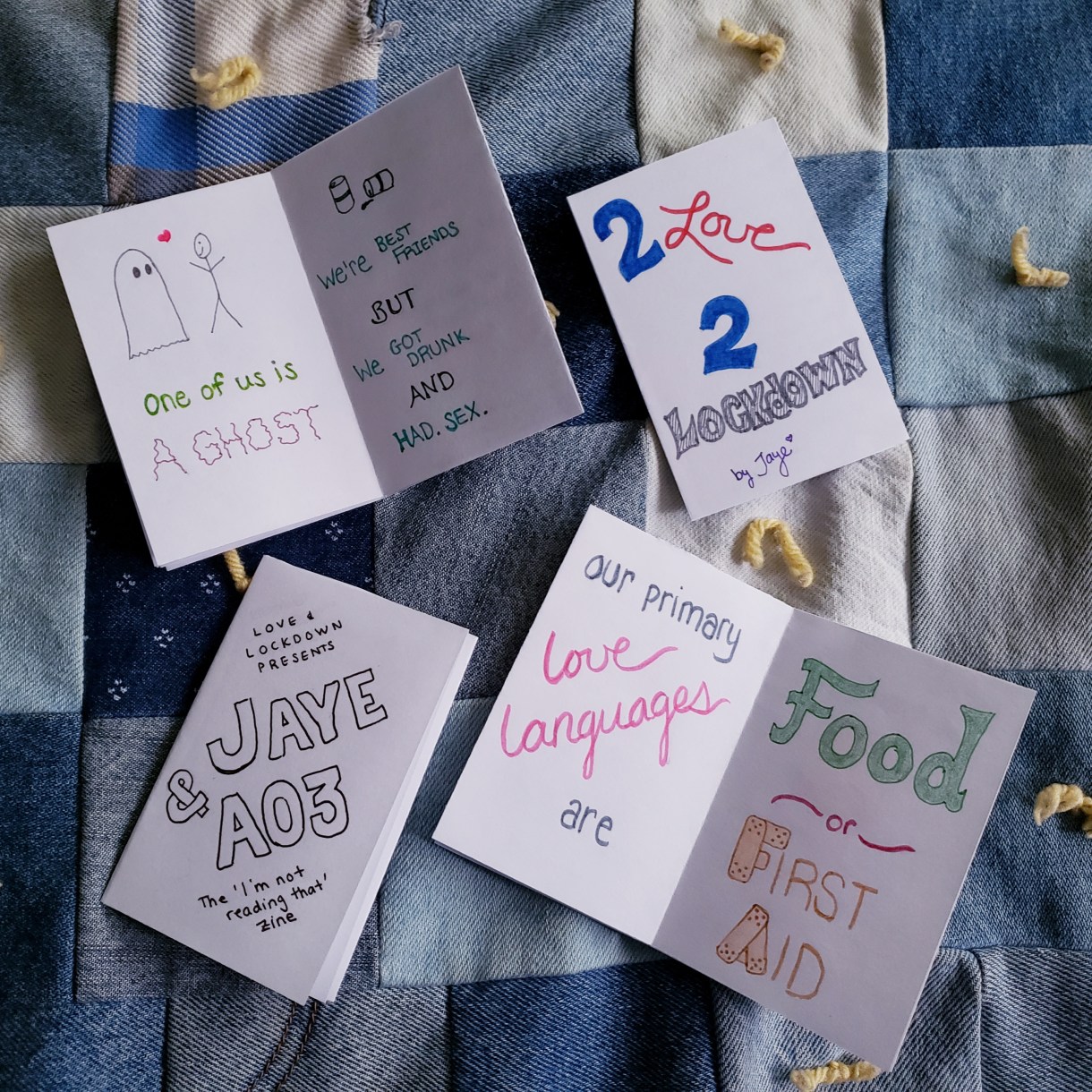 There are four zines laying on a quilted background. One reads: "One of us is a Ghost. We're best friends but we got drunk and had sex" one reads: 2 Love 2 Lockdown, another says "Love and Lockdown presents Jayea & A03, The I'm Not Reading That Zine" and finally, an open zine that reads "Our primary love languages are food or first aid."