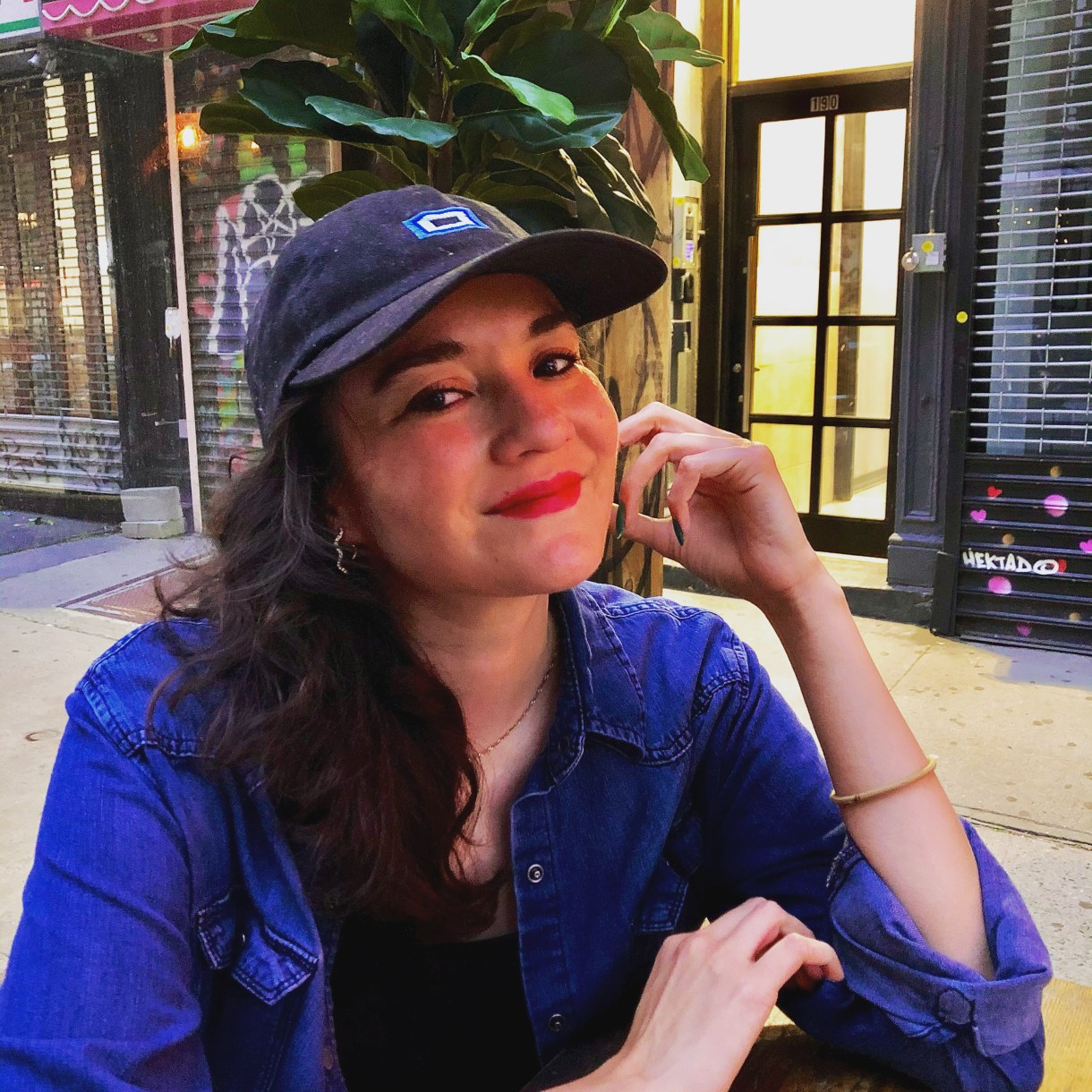 Analyssa is a young latine woman here, wearing a baseball cap and denim buttom up shirt with a hairband, long nails painted a dark color and lipstick. She also has long dark brown hair with a slight wave and brown eyes. She is giving the camera a knowing smile.