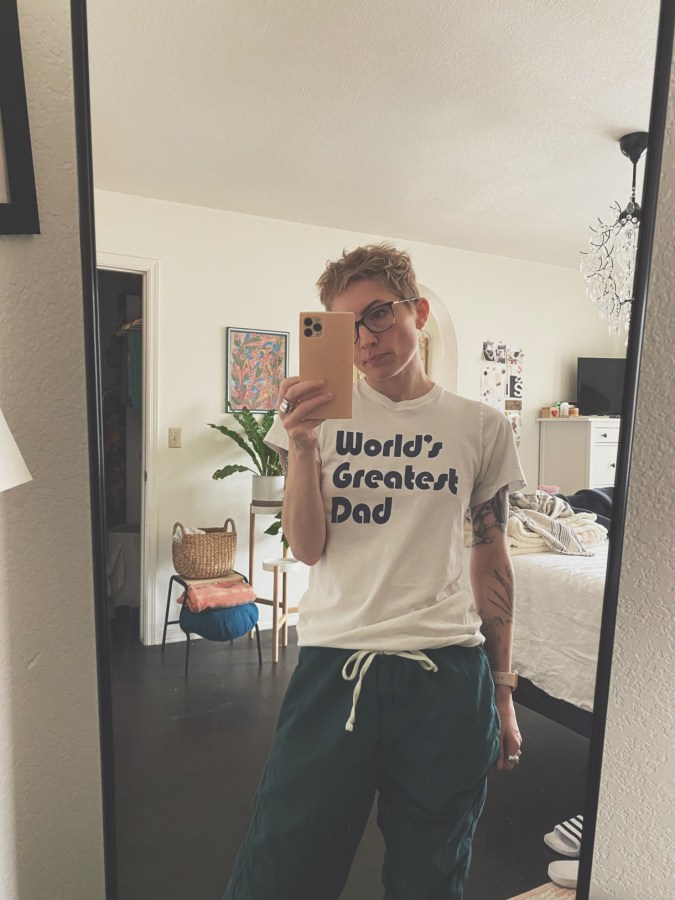 Laneia, a white woman with short bleached hair and glasses, is wearing a "World's Greatest Dad" tee