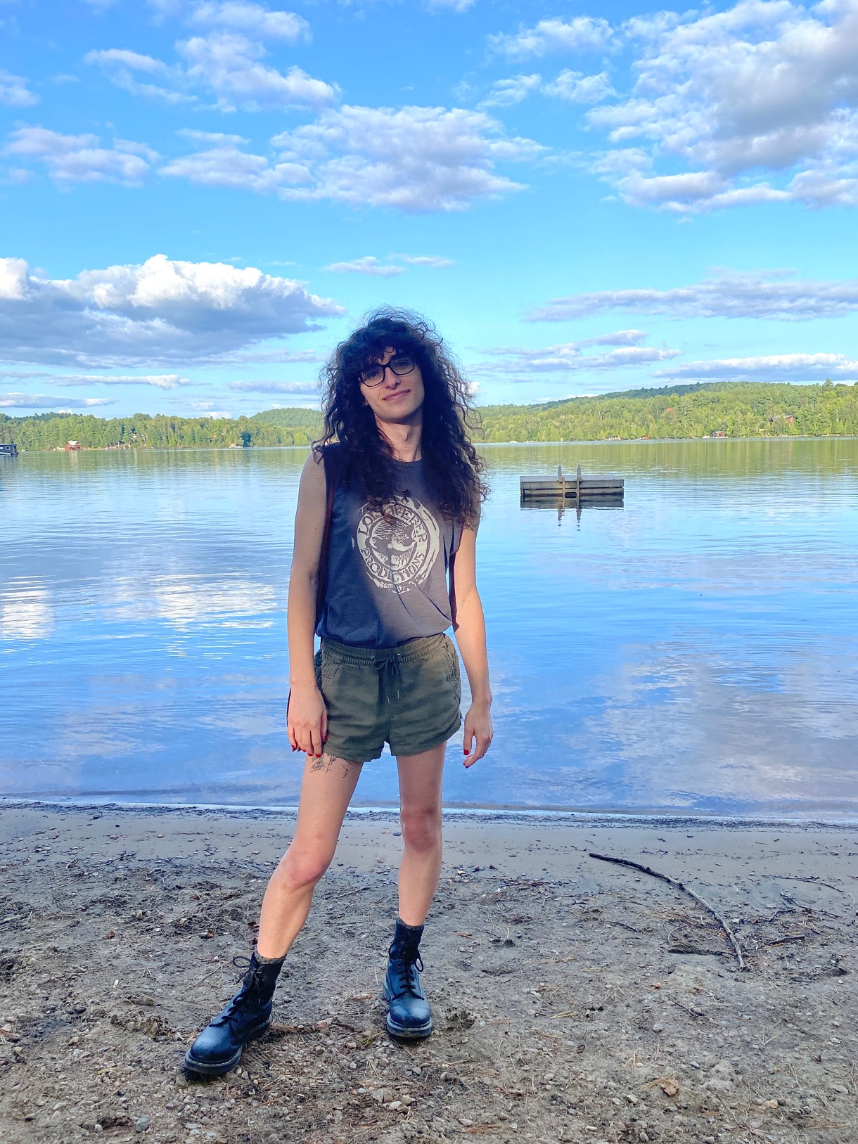 Drew, a white woman with long curly brown hair and glasses, is wearing a Lois Weber productions tee