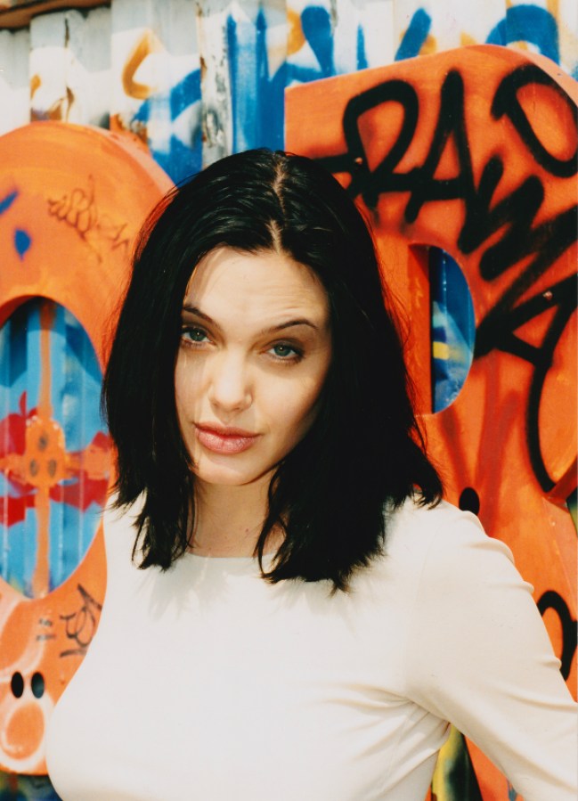 Angelina Jolie stands in front of an orange graffiti wall with dark black hair that is shoulder length and a white sweater.