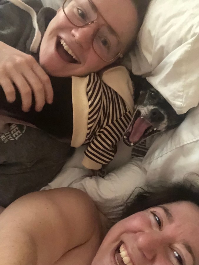two dykes in bed snuggling a tiny chihuahua with his mouth open