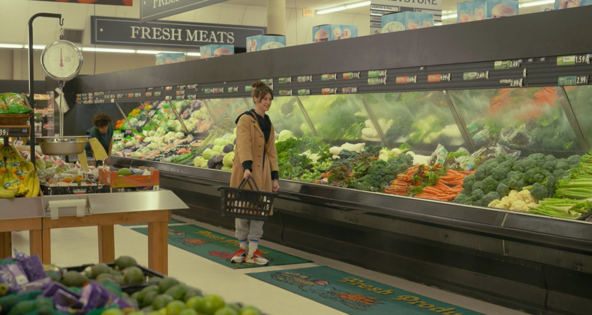 Daisy Edgar-Jones wearing a camel colored coat and lightwash jeans while holding a grocery basket in the produce section of a grocery store and standing under a sign that says Fresh Meats