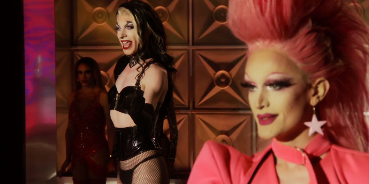 Drag Race recap 1411: In the foreground Lady Camden with big pink hair and a pink star earring is out of focus, in the background Bosco in black leather clenches her fist while belting