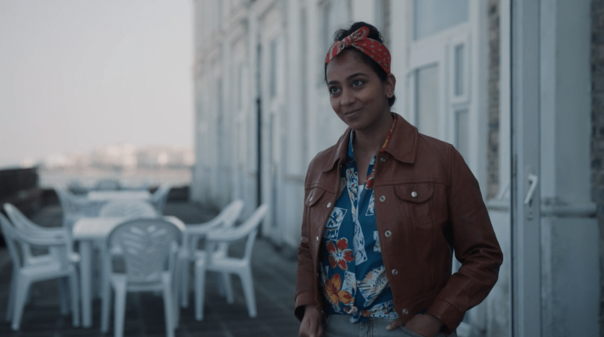 Pam wears a light brown leather jacket over a patterned buttondown on Killing Eve. She is smiling and also wearing a red bandana in her hair, which is pulled back.