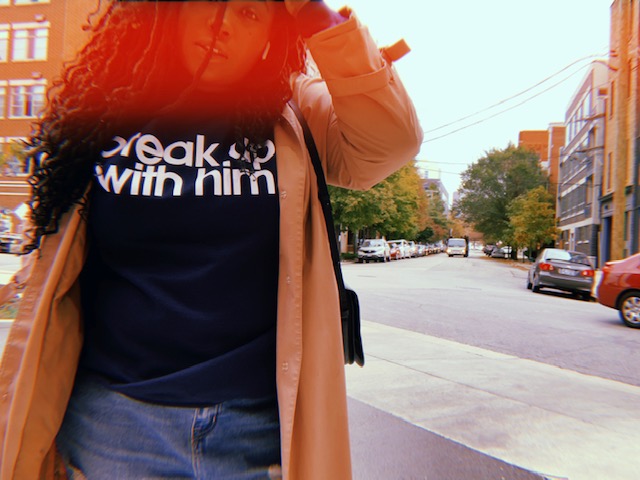 Shelli, a Black woman with long hair is wearing a camel coat over a tee that reads "Break Up With Him"