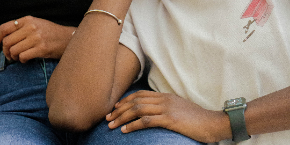 The image shows the chest and arms of a Black woman wearing a white T-shirt with a graphic of an open cigar box on one side of the chest, a gray watch on one wrist and a simple silver bracelet on the other wrist. She is leaning against another woman, who was wearing a black shirt and jeans. Only one of her hands and her upper legs are visible.