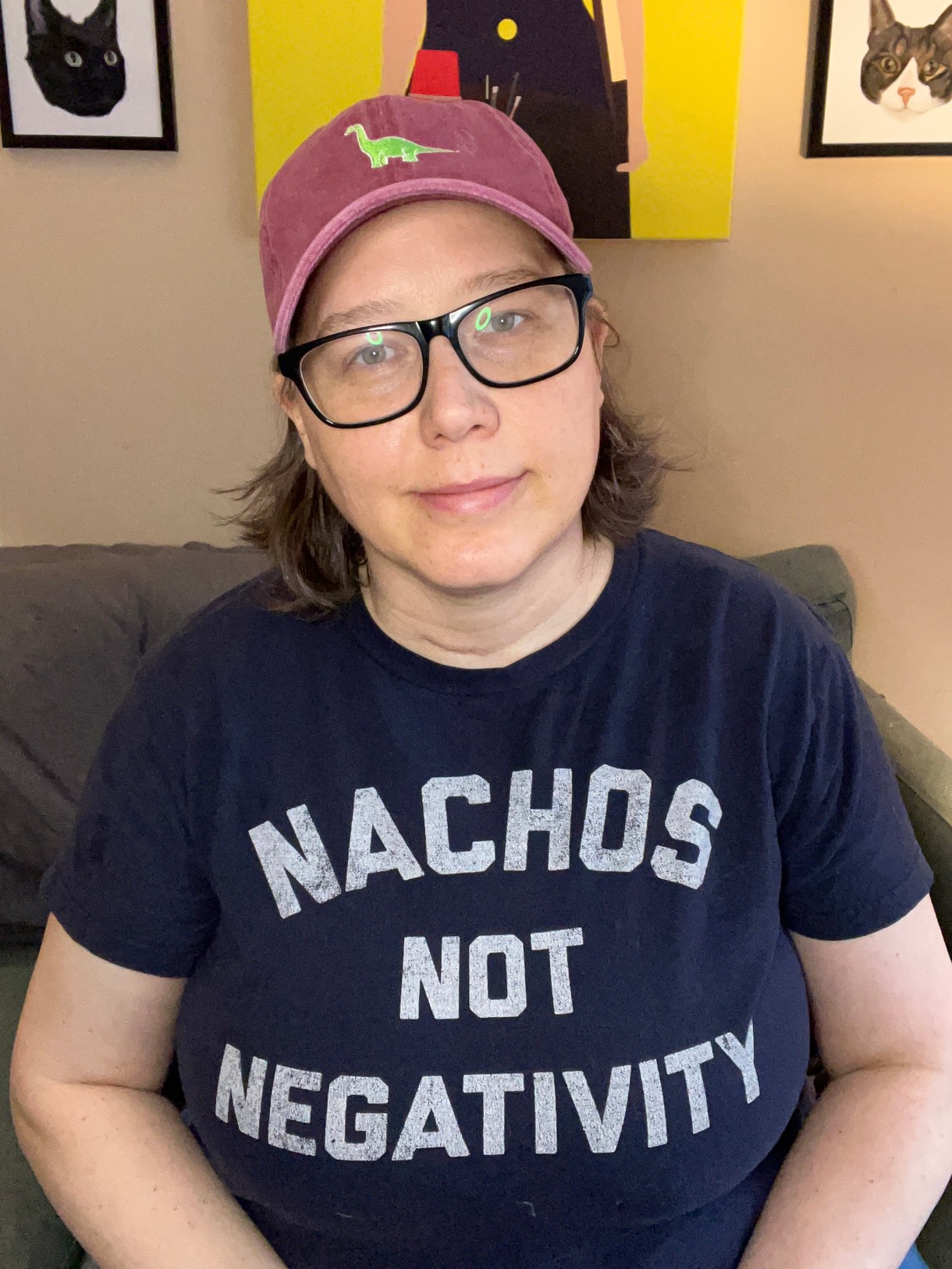 Heather, a white woman with short blonde and gray hair, glasses and a baseball cap is wearing a shirt that says "Nachos Not Negativitiy"