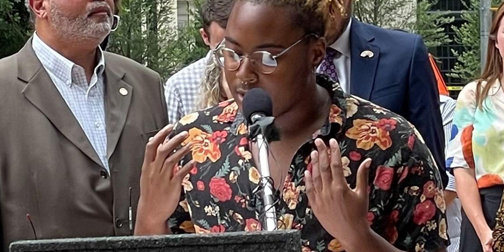 A dark-skinned Black masculine person is wearing a colorful shirt and speaking into a mic at a podium.