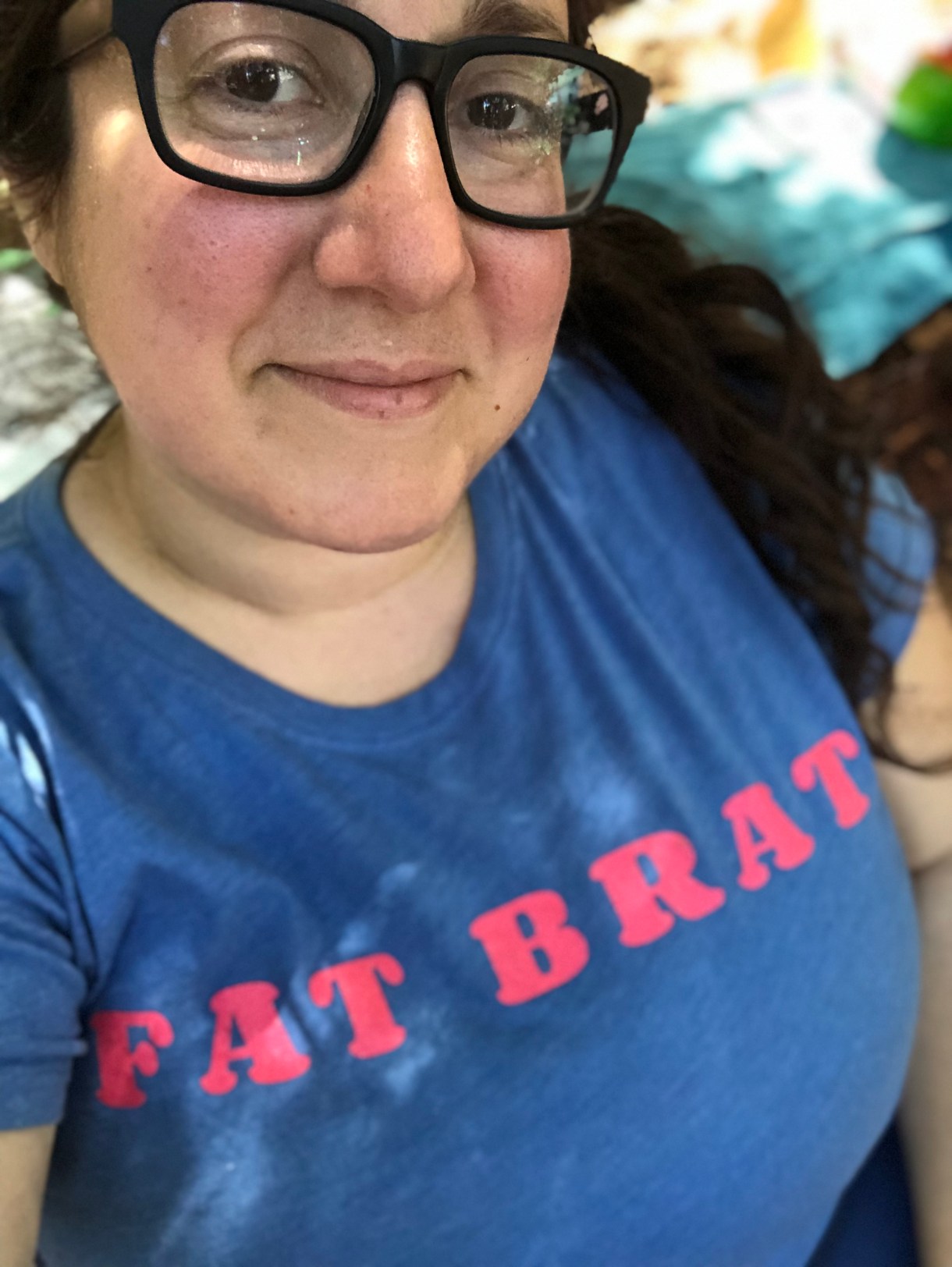 Vanessa, a white woman with long brown hair and glasses is wearing a shirt that reads "Fat Brat"