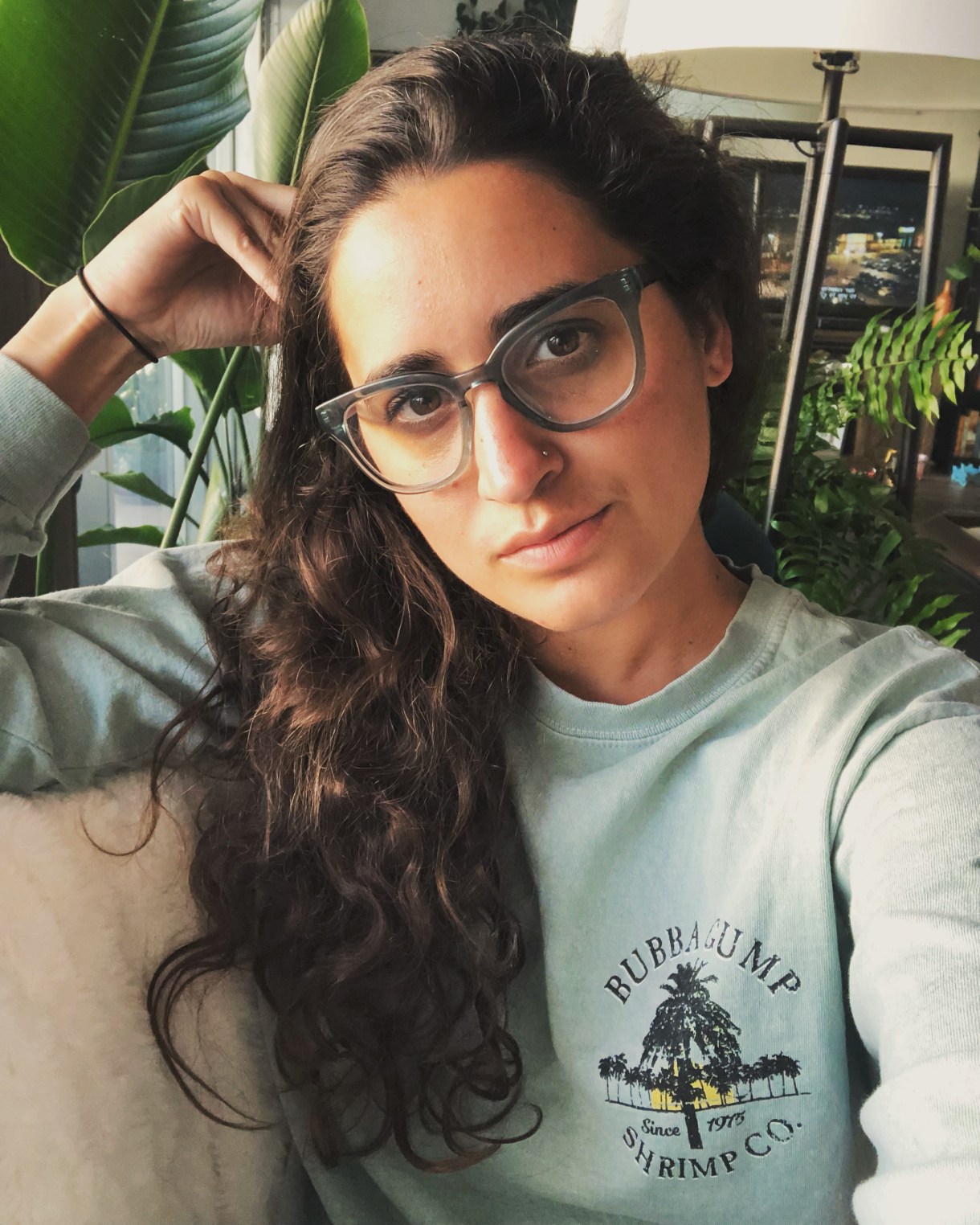 Kayla Kumari Upadhyaya wears blue framed glasses and a long sleeve pale green t-shirt that says Bubba Gump Shrimp Co. Since 1975 on it.