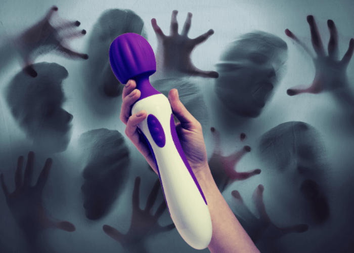 a pale arm holds up magic wand style vibrator in front of a background of ghostly figures pressing through a translucent background. the background has a cheap,  spooky haunted house feel