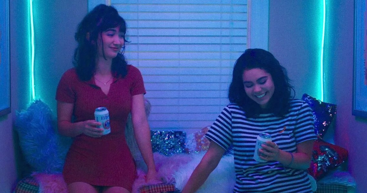 In a still from Hulu's crush, Rowan Blanchard's Paige is smiling while facing right of camera, she is making Auli'i Cravalho's AJ blush. The lighting is shades of blue and purple. Paige is in a red dress with brunette hair half up in a pony tail. AJ is in a black and white striped shirt with brunette wavy hair in a messy bob.