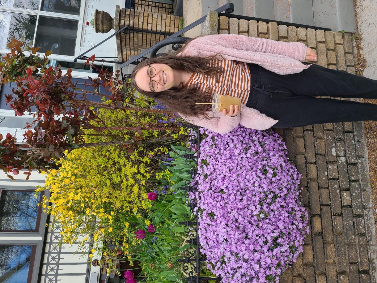Anya, a white woman with glasses and long brown hair, stands with an iced coffee drink in front of a house with flowers in front