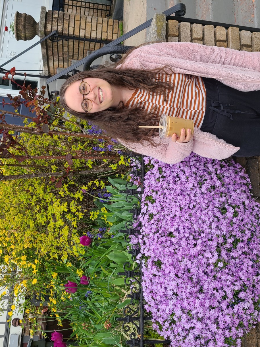 Anya, a white woman with glasses and long brown hair, stands with an iced coffee drink in front of a house with flowers in front