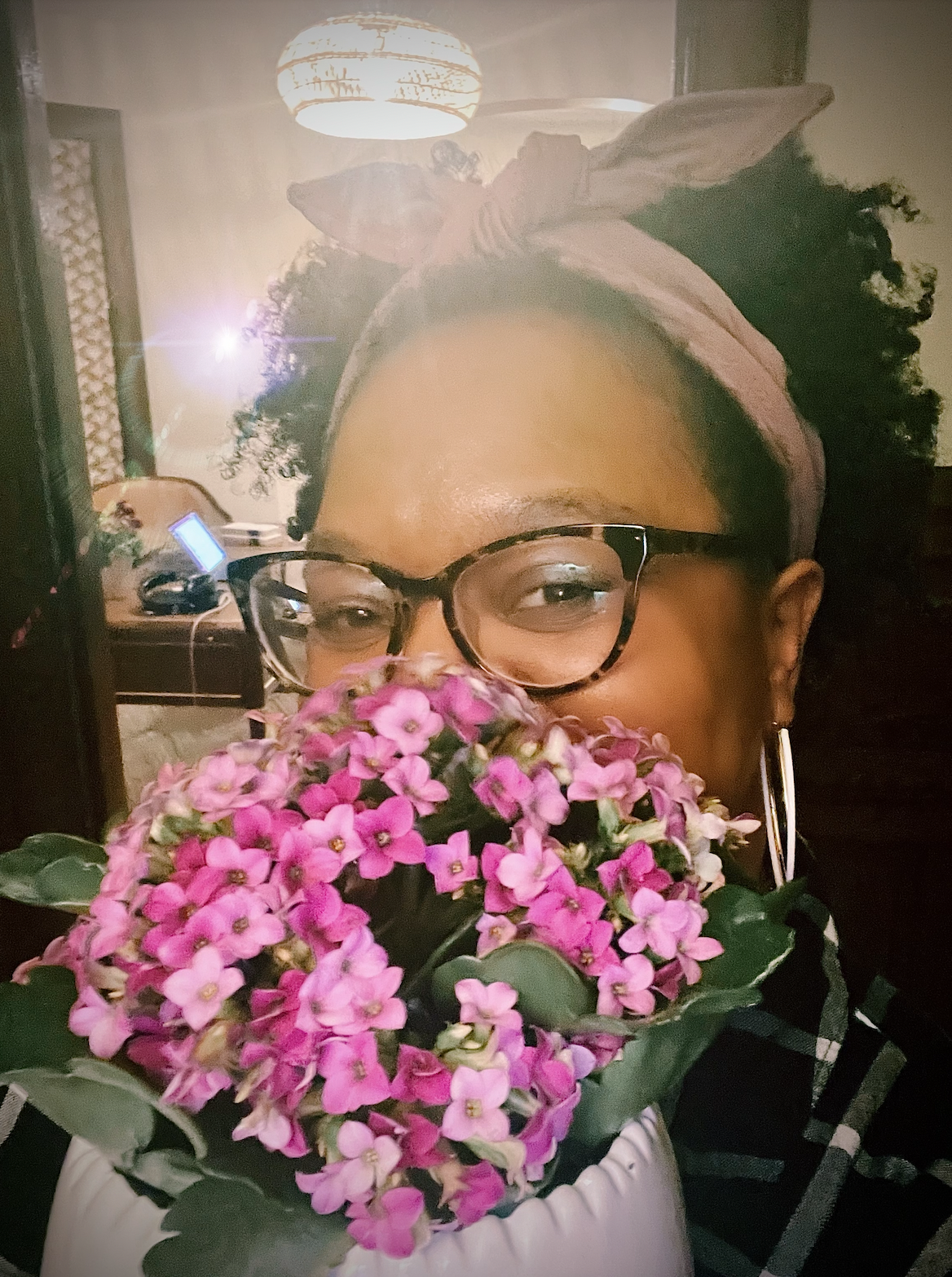 Carmen, a Black woman wearing glasses and hoop earrings and a pink scarf tied up at the top of her head. Her curly hair is parted in the middle. She is smiling with her eyes over the top of some pink flowers.