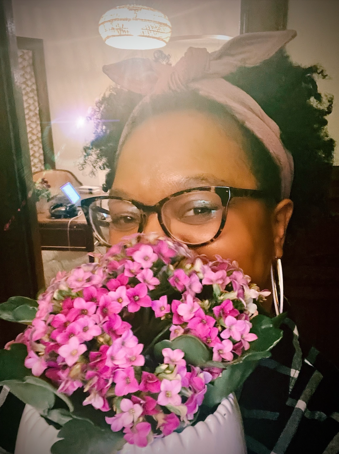 Carmen, a Black woman wearing glasses and hoop earrings and a pink scarf tied up at the top of her head. Her curly hair is parted in the middle. She is smiling with her eyes over the top of some pink flowers.