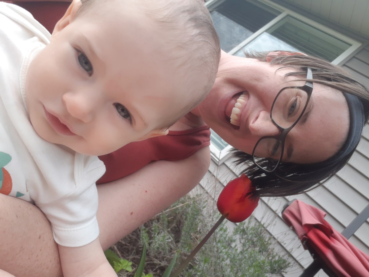 Casey with her baby! Casey is a white woman with short brown hair and glasses. They are laughing in front of a single red tulip.