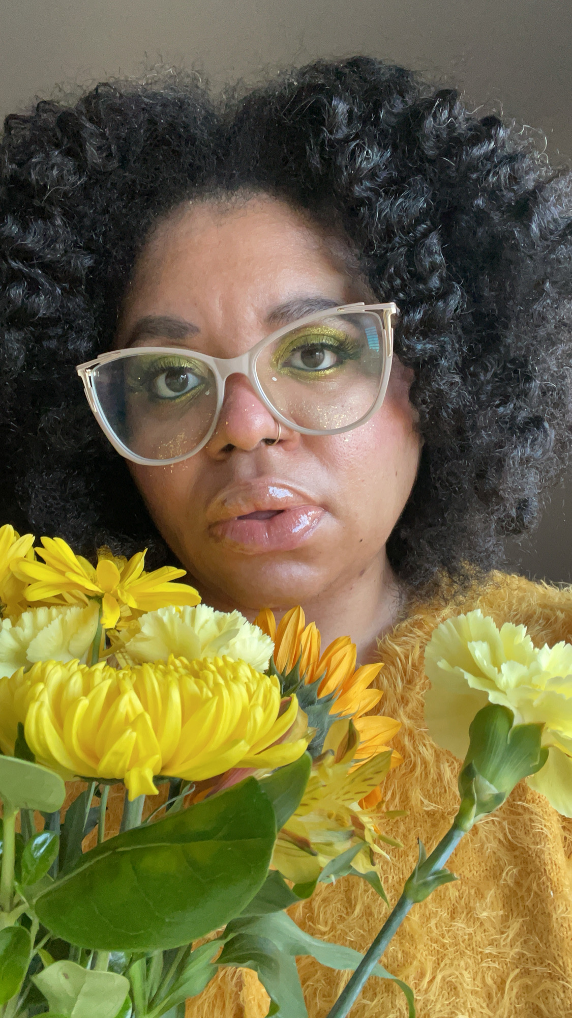 Dani is a Black woman with shiny, loosely curled hair and large vintage-inspired glasses. She is gazing at the viewer and holding a bouquet of gold, yellow and orange flowers and is wearing a matching gold top.