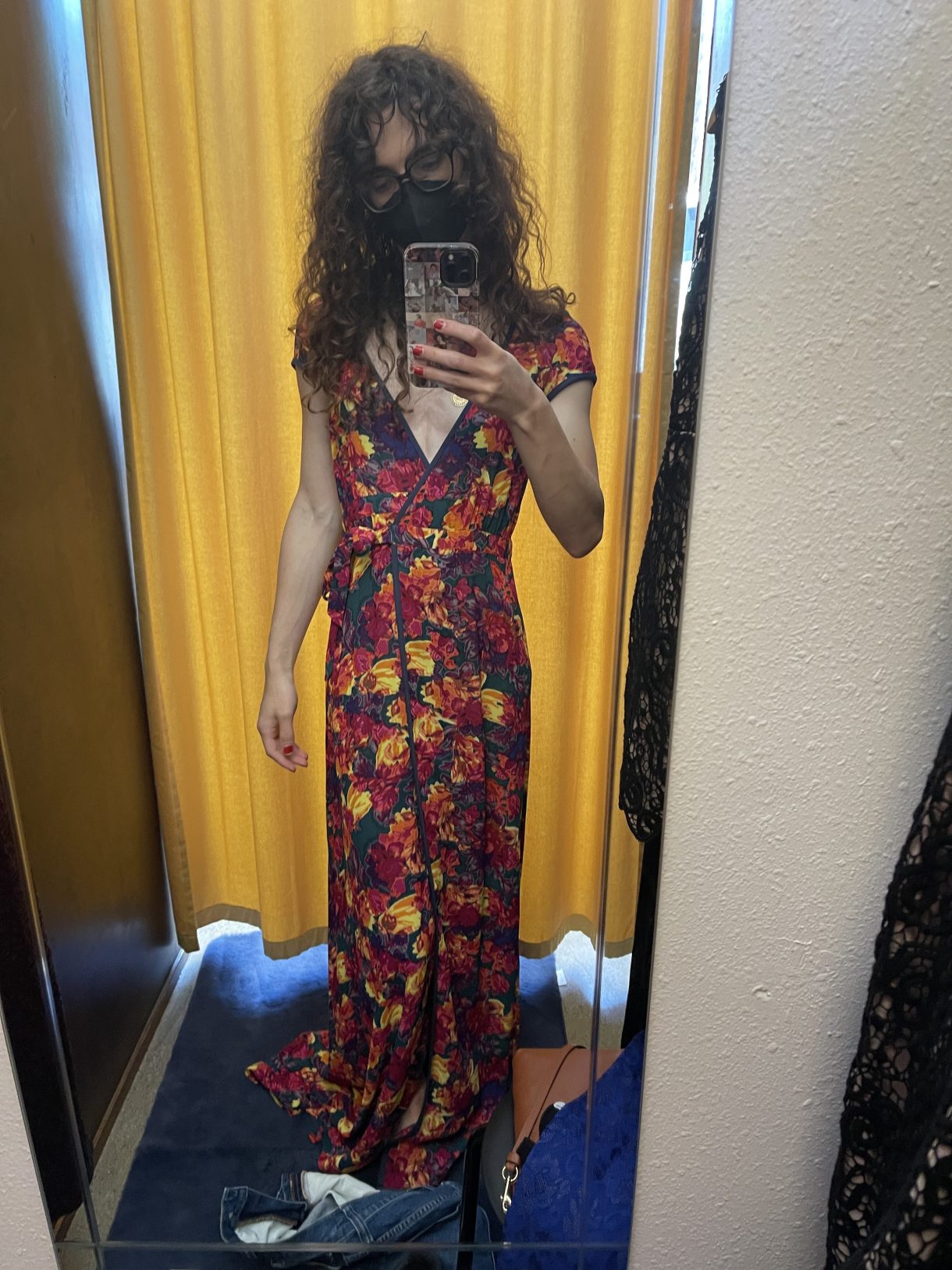 Drew is a white woman with long, curly brown hair. She is wearing a floor-length floral dress in vibrant colors and appears to be in a changing room.