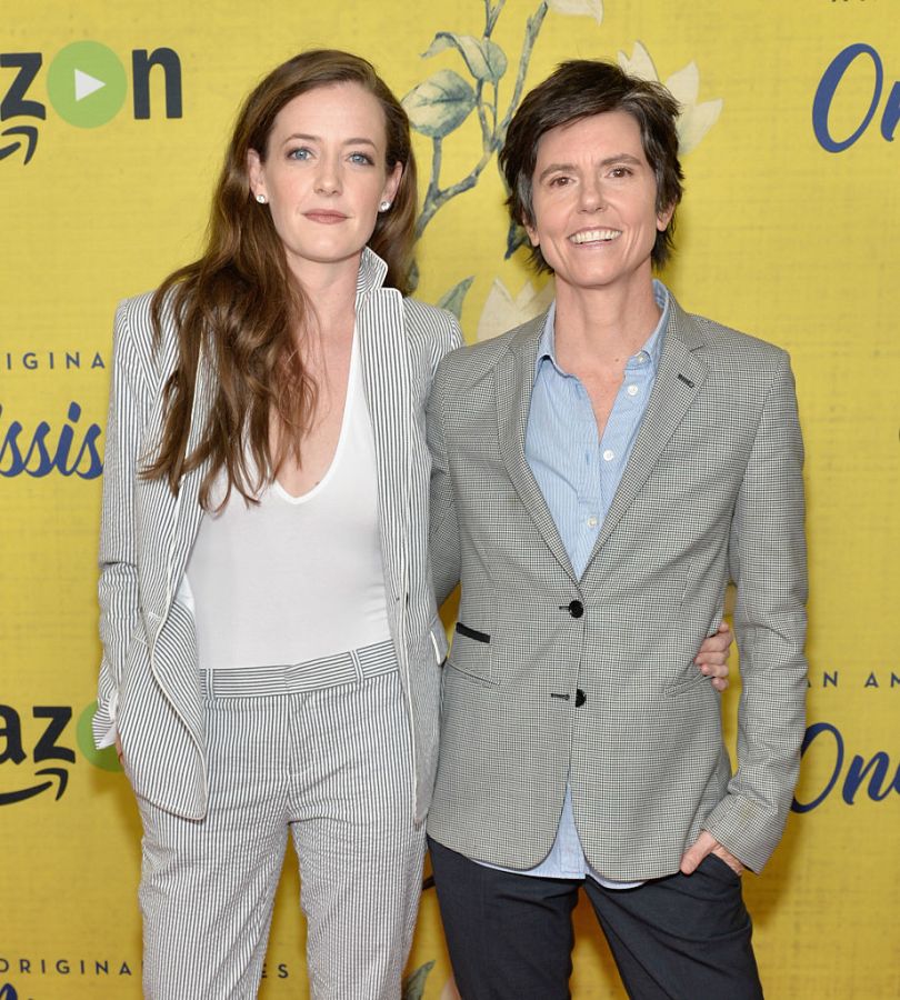 Stephanie Allynne and Tig Notaro attend the premiere of Amazon Instant Video's 'One Mississippi' at The London West Hollywood. Stephanie is wearing a white shirt with a striped blazer and matching pants. Tig is wearing a blue buttondown and a gray blazer. They have their arms around each other and are smiling at the camera.