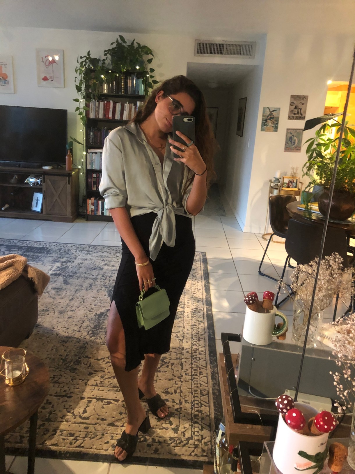 Kayla Kumari Upadhyaya wears a pale blue silky buttondown blouse that's tied at the waist and a tight black long skirt with a slit at the thigh. She is holding a tiny green purse and also wearing low-heeled black mules.