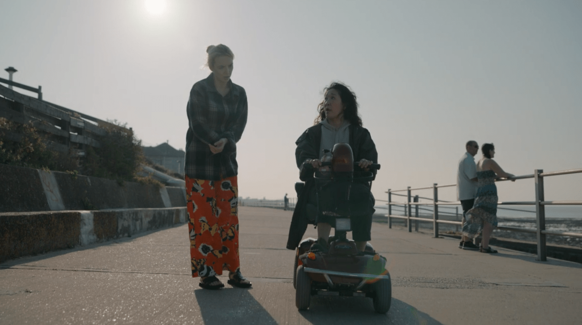 Villanelle wears a flannel over a patterned jumpsuit. Next to her, Eve is on a mobility scooter in a hoodie.