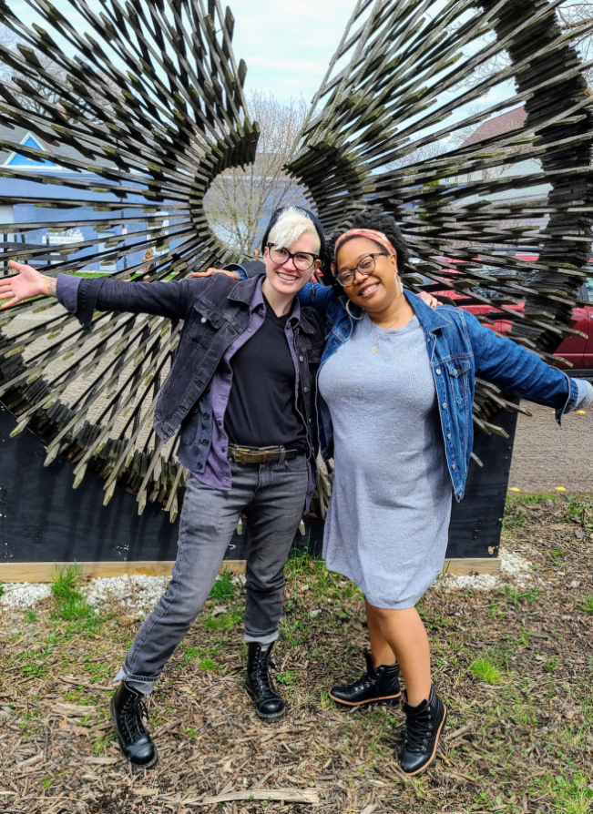 Nicole and Carmen pose arm in arm, with outside arms outsretched, smiling in front of a sculpture outdoors. Carmen is a Black woman who is wearing a headband tied in a bow at the top, and who has her curly hair parted in the center. She is wearing a jean jacket over a sweater dress and black boots that come up over the ankle as well as glasses and hoop earrings. Nicole is a white genderqueer human who is wearing a black jean jacket, gray jeans, a v neck black tee, a couple of random gray flannel layers and black combat boots along with a beanie and glasses. They are both smiling sweetly for the camera!
