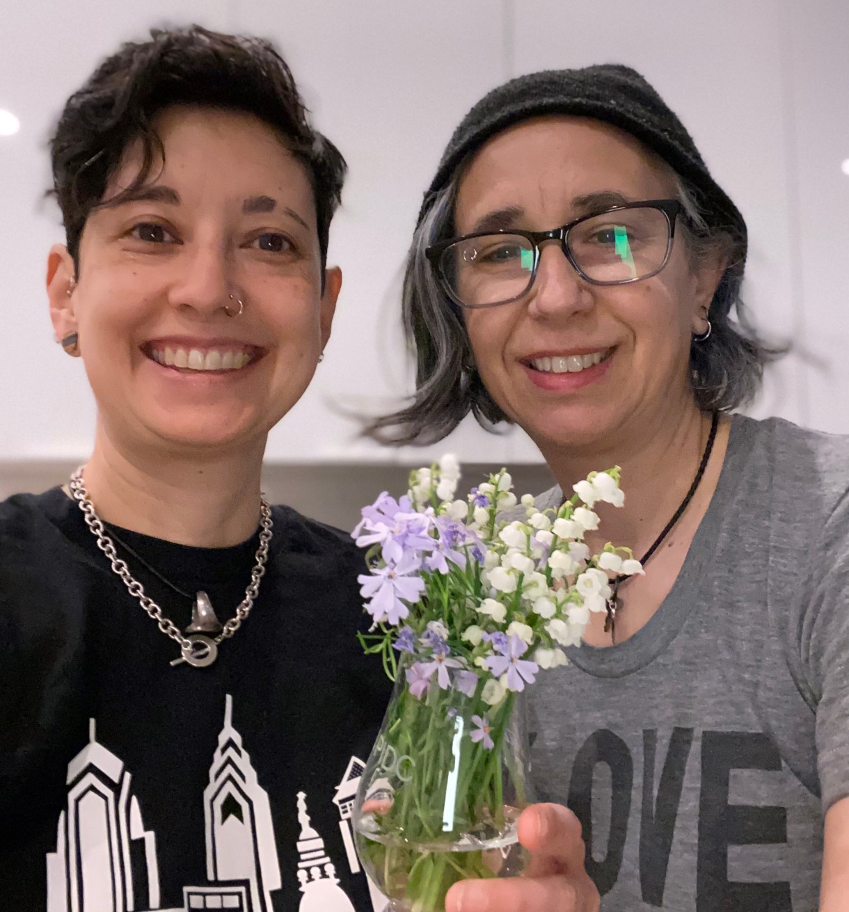 Tracy and Mia from Yikes our tech team hold up a vase of beautiful little flowers. Tracy is an Asian woman with short black hair and a nose piercing and Mia is a white woman with gray hair  who is wearing glasses and a beanie.