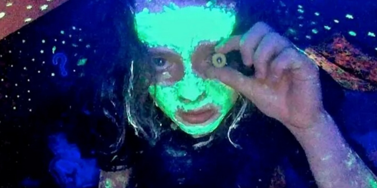 Casey, played by Anna Cobb, looks into her webcam with paint on her face that shines green in her blacklight. She holds a fake eye in front of her left eye in Jane Schoenbrun's movie We're All Going to the World's Fair.